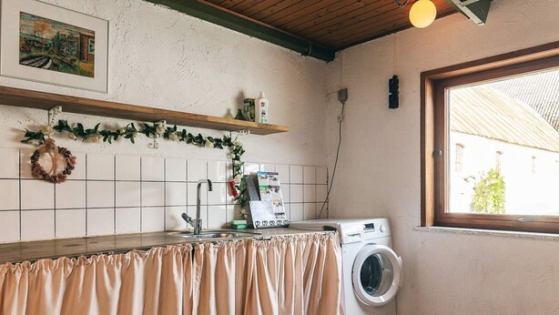 A laundry area with a washing machine, a sink, a shelf with a wreath, and a window with a view of buildings. - 4 Star Holiday Home in Aabybro (Aabybro)