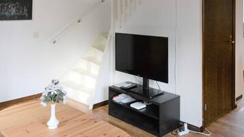 Una sala de estar con un televisor de pantalla plana sobre un soporte negro, una mesa de madera con un jarrón blanco y una escalera al fondo.