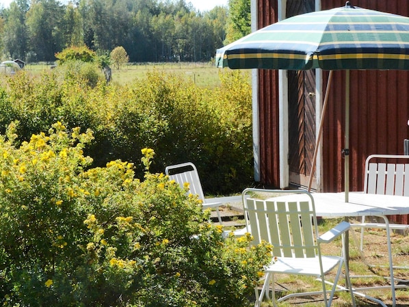 An outdoor seating area with a table and chairs, a striped umbrella, and a view of a field and trees.