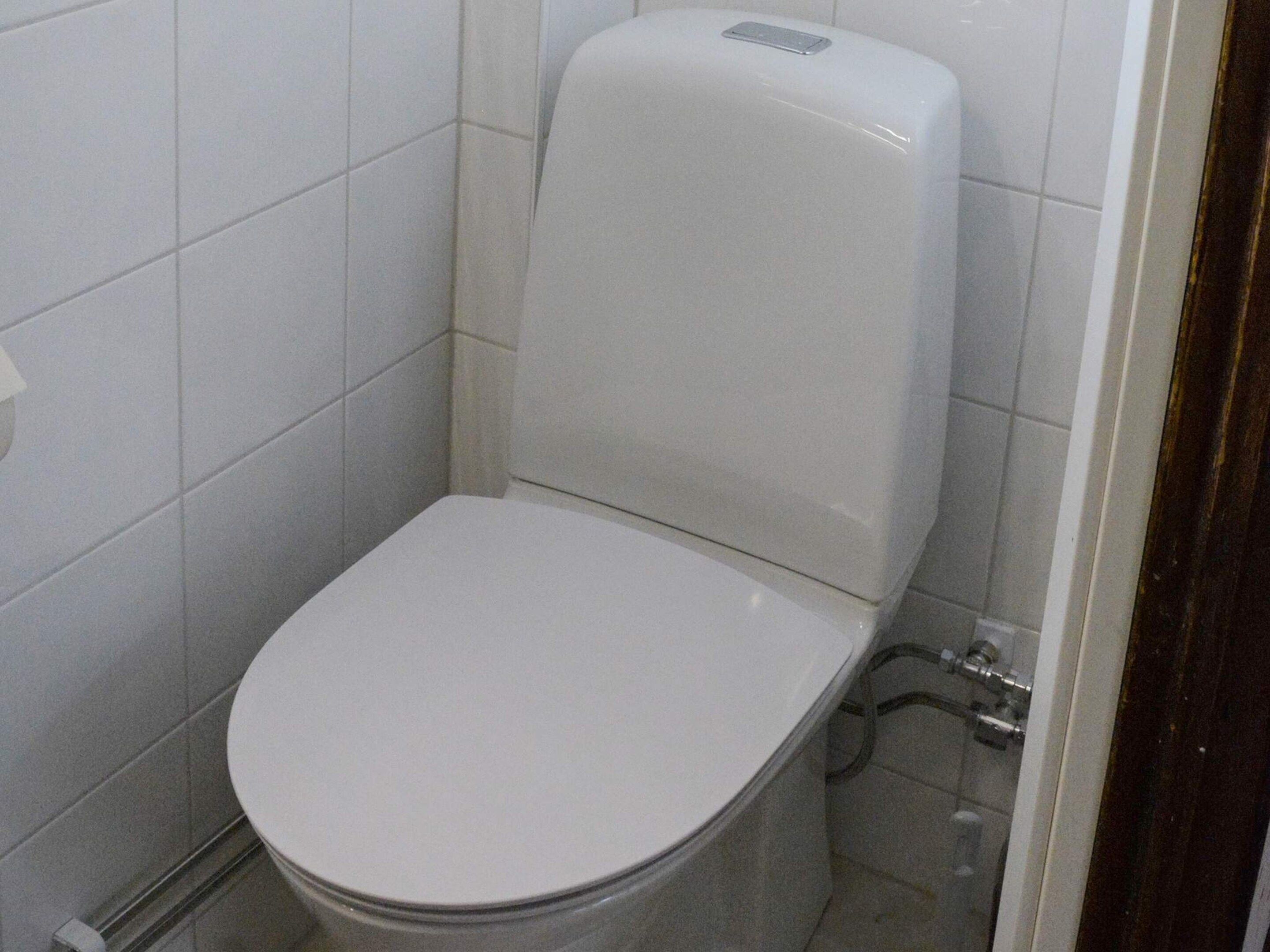 Et standard hvid toilet i et badeværelse med fliserede vægge og synlige VVS-installationer.