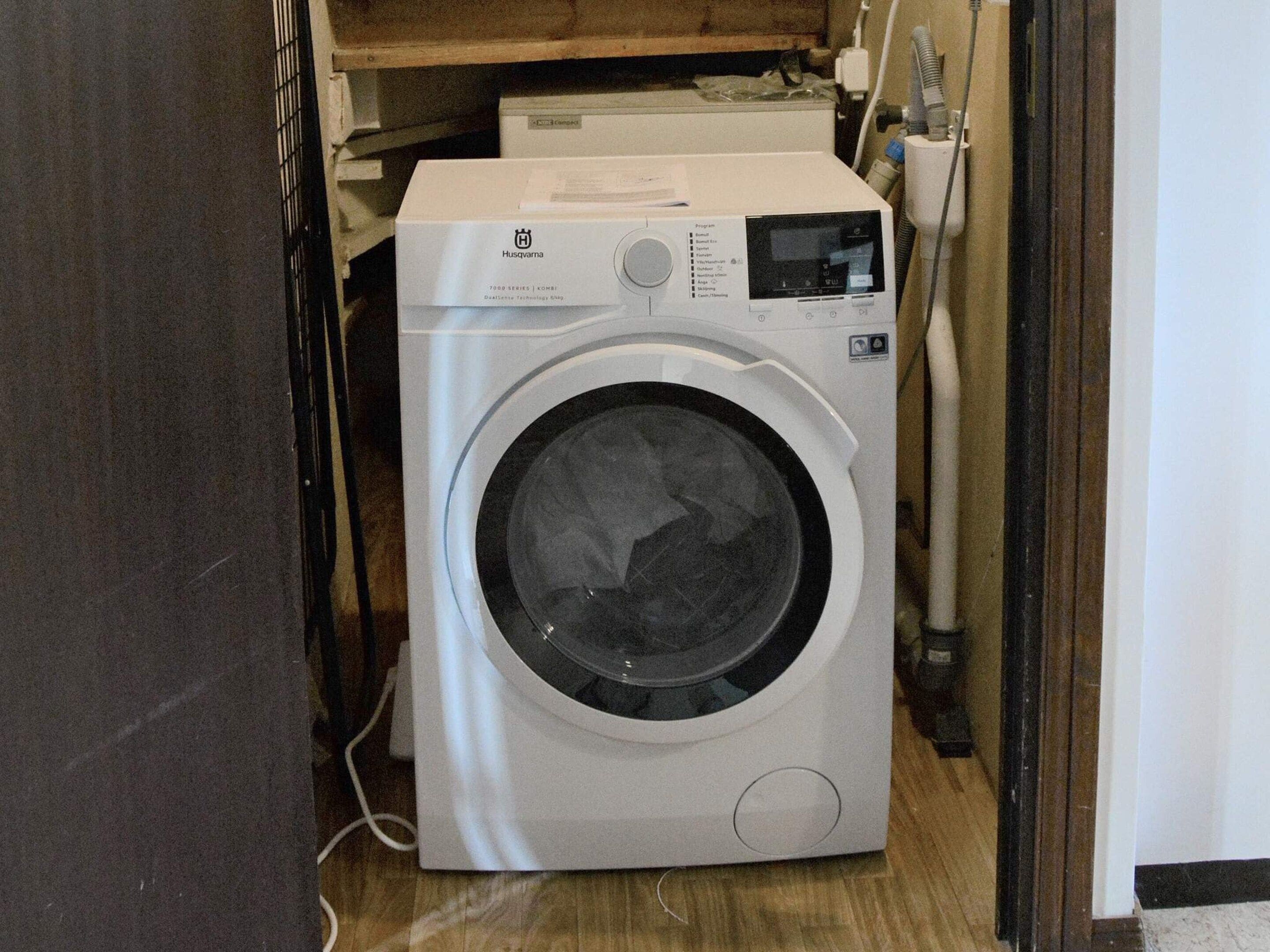 A front-loading washing machine in a laundry room with a wooden floor and a white wall.