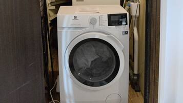 A front-loading washing machine in a laundry room with a wooden floor and a white wall.