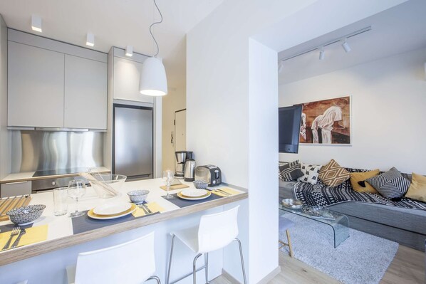 A modern kitchen with a central island, a dining area, and a living space with a sofa and a painting. - Alterra Vita Thessaloniki Apartment (Thessaloniki)