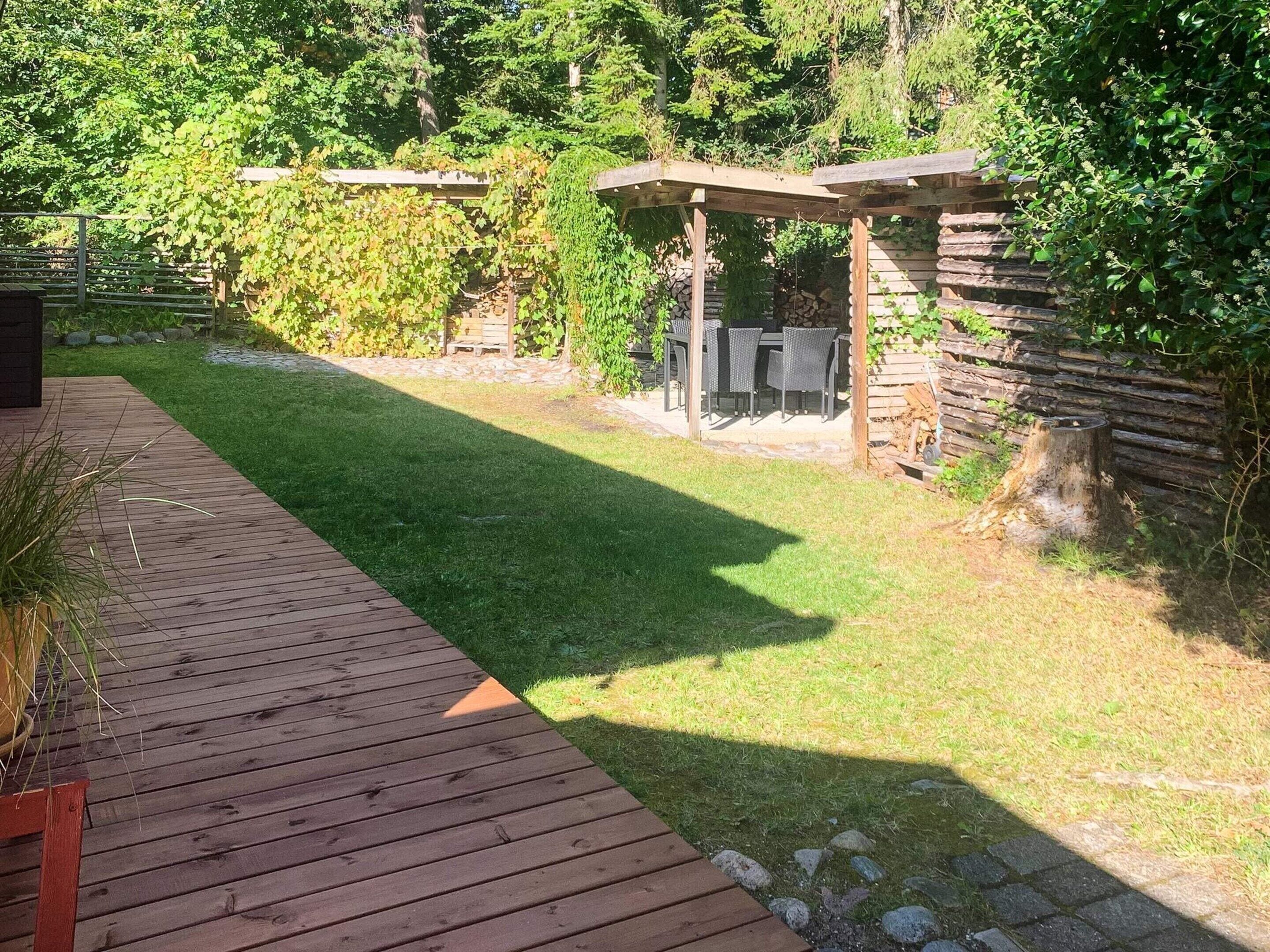 Une terrasse en bois mène à un espace extérieur aménagé, abrité, avec un mur en pierre et de la verdure en arrière-plan.