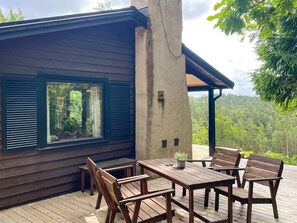 Une terrasse en bois avec une table et des chaises, une fenêtre avec des volets et une vue sur une zone boisée.