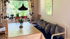 A room with a dining table, a sofa with patterned cushions, a hanging light fixture, and a window with curtains.