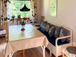 Ett rum med ett matbord, en soffa med mönstrade kuddar, en hängande lampa och ett fönster med gardiner.