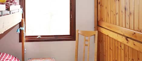 A room with a bunk bed, a small wooden chair, a window with a view, and a wooden wardrobe.