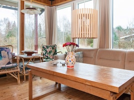 A wooden dining table with a vase of flowers, a wicker chair, and a sofa in a room with large windows and a hanging light fixture.
