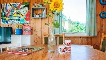A wooden interior with a dining table, a vase with a sunflower, a mug, and a colorful painting.