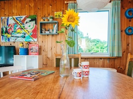 A wooden interior with a dining table, a vase with a sunflower, a mug, and a colorful painting.