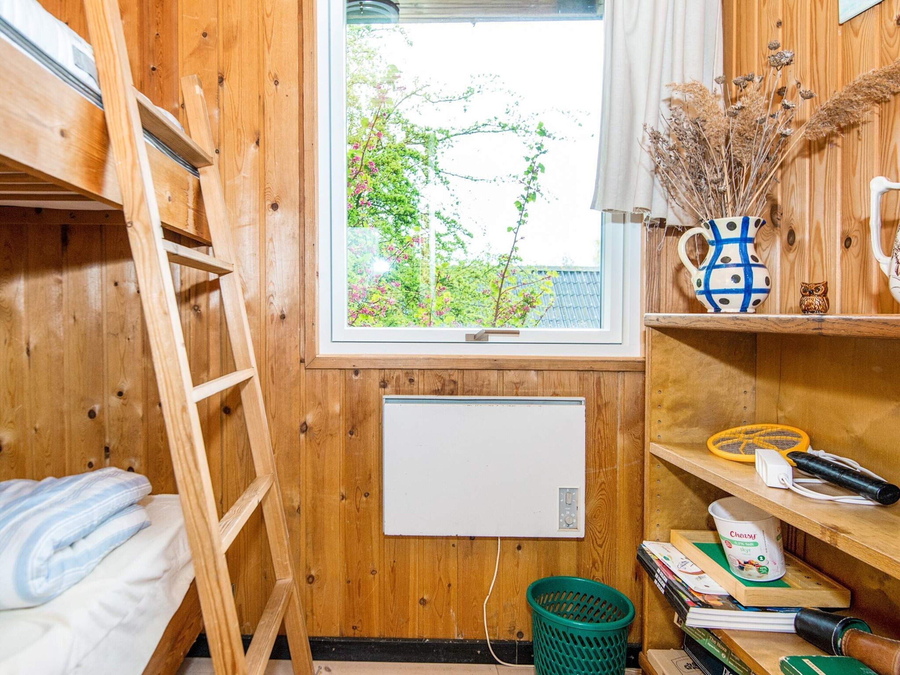 A wooden room with bunk beds, a window with a view of greenery, a radiator, and shelves with various items.