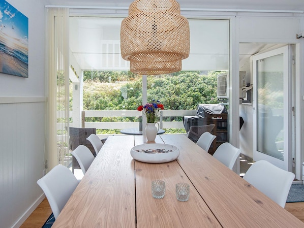Un comedor con mesa de madera, sillas blancas, una lámpara colgante de fibra y vistas a zonas verdes a través de amplios ventanales.