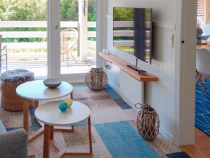 Ein modernes Wohnzimmer mit an der Wand montiertem Fernseher, einem Holztisch, zwei Stühlen und Blick ins Freie durch große Glasüren.