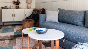 Ein Wohnzimmer mit einem blauen Sofa, einem weißen Tisch mit einer Schale, einem weißen Schrank, einer Lampe und einem gerahmten Bild an der Wand.