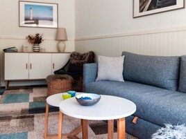 Una sala de estar con un sofá azul, una mesa blanca con un cuenco, un armario blanco, una lámpara y un cuadro en la pared.