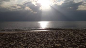 Una playa con huellas que conducen al agua, cielo despejado y el sol brillando entre las nubes.