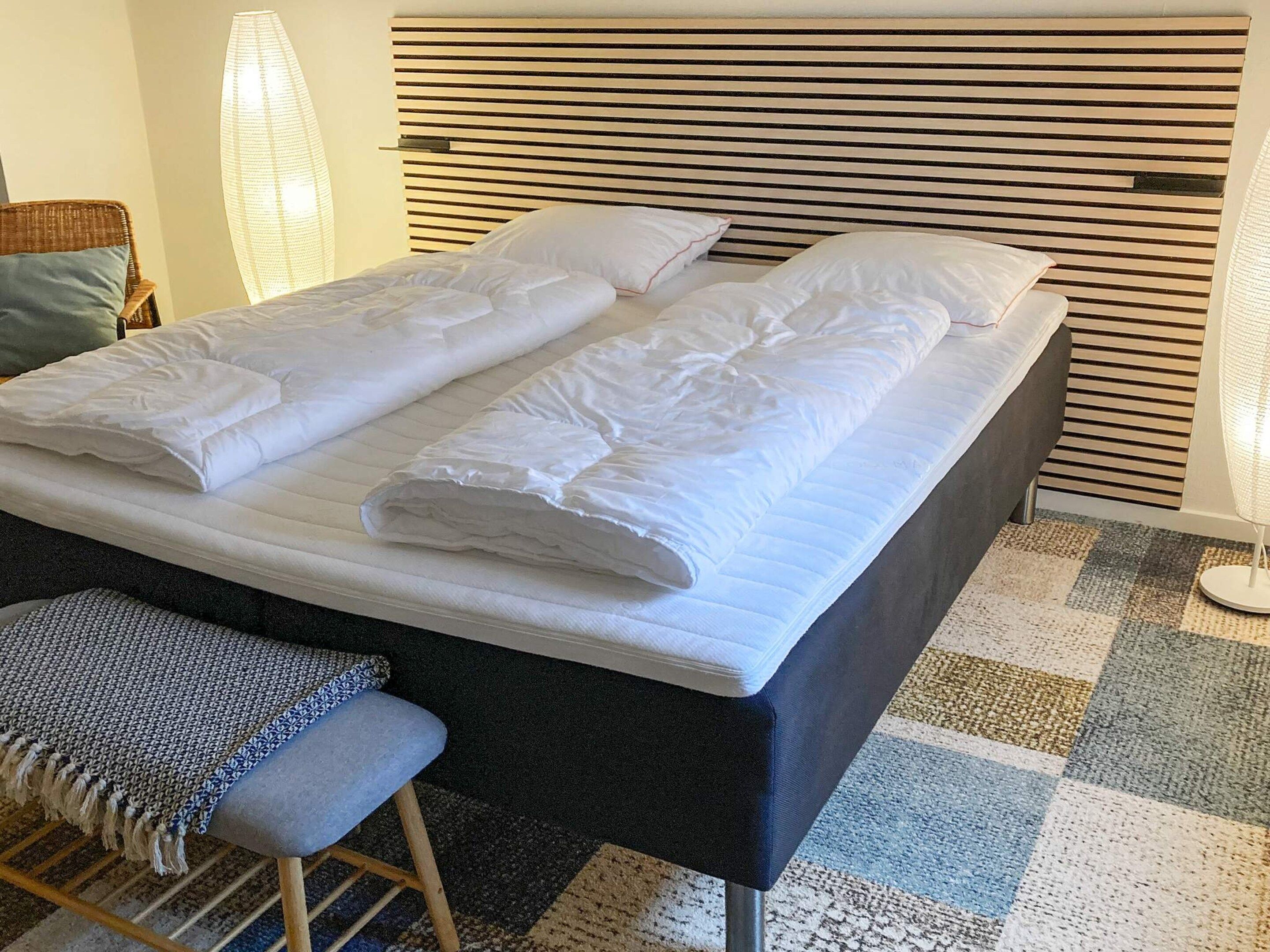 Una cama bien hecha con almohadas y un edredón doblado, un banco con cojín, una alfombra estampada y un cabecero moderno.