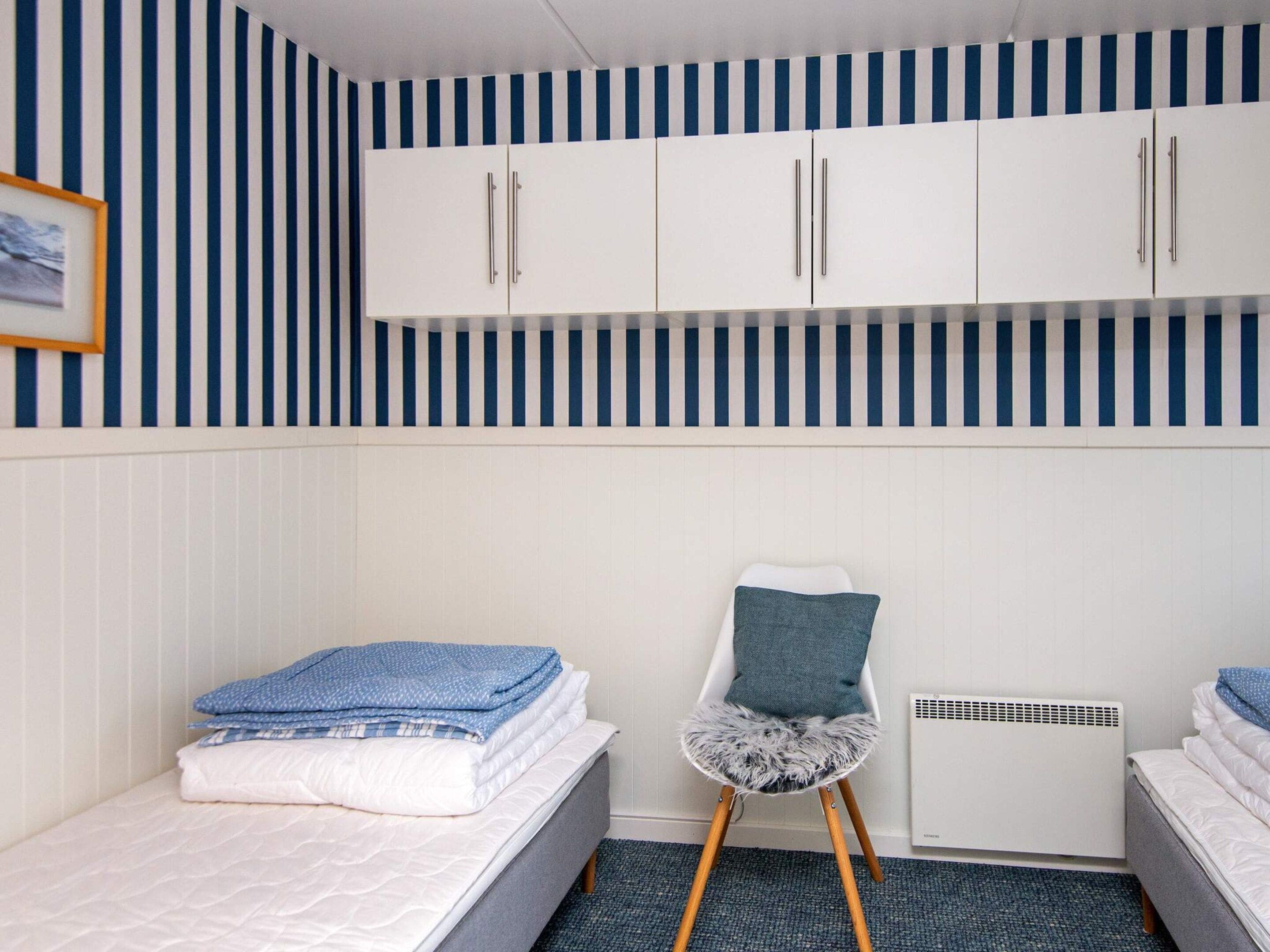 A room with a striped wall, a bed with folded blankets, a chair with a cushion, and a white cabinet.