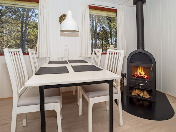 A dining area with a white table, black placemats, white chairs, a white pendant light, and a wood-burning stove.