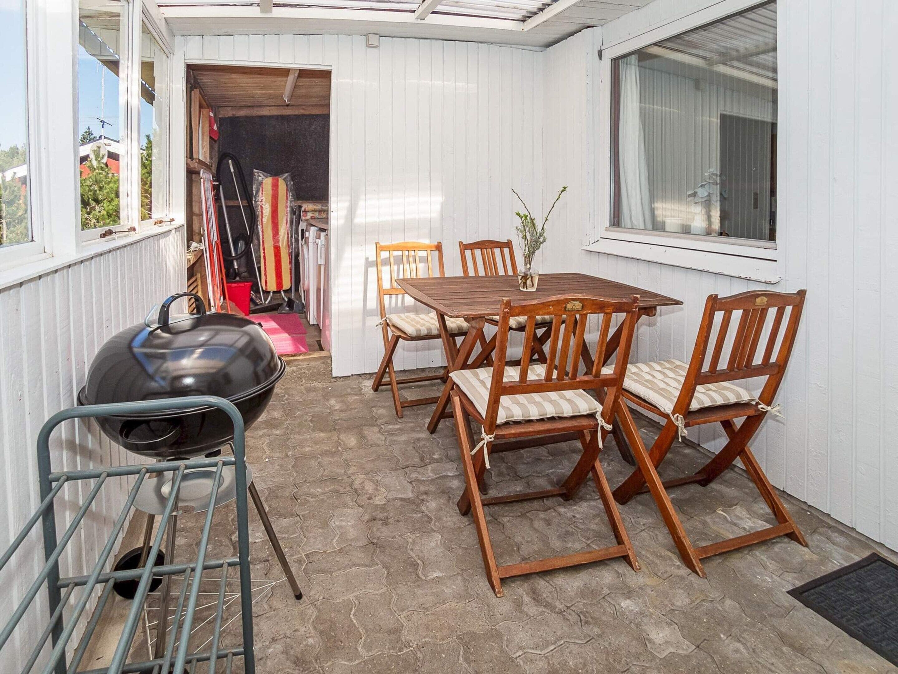 Eine Terrasse im Freien mit einem Holztisch und Stühlen, einem Grill und einem Fenster mit Vorhängen.