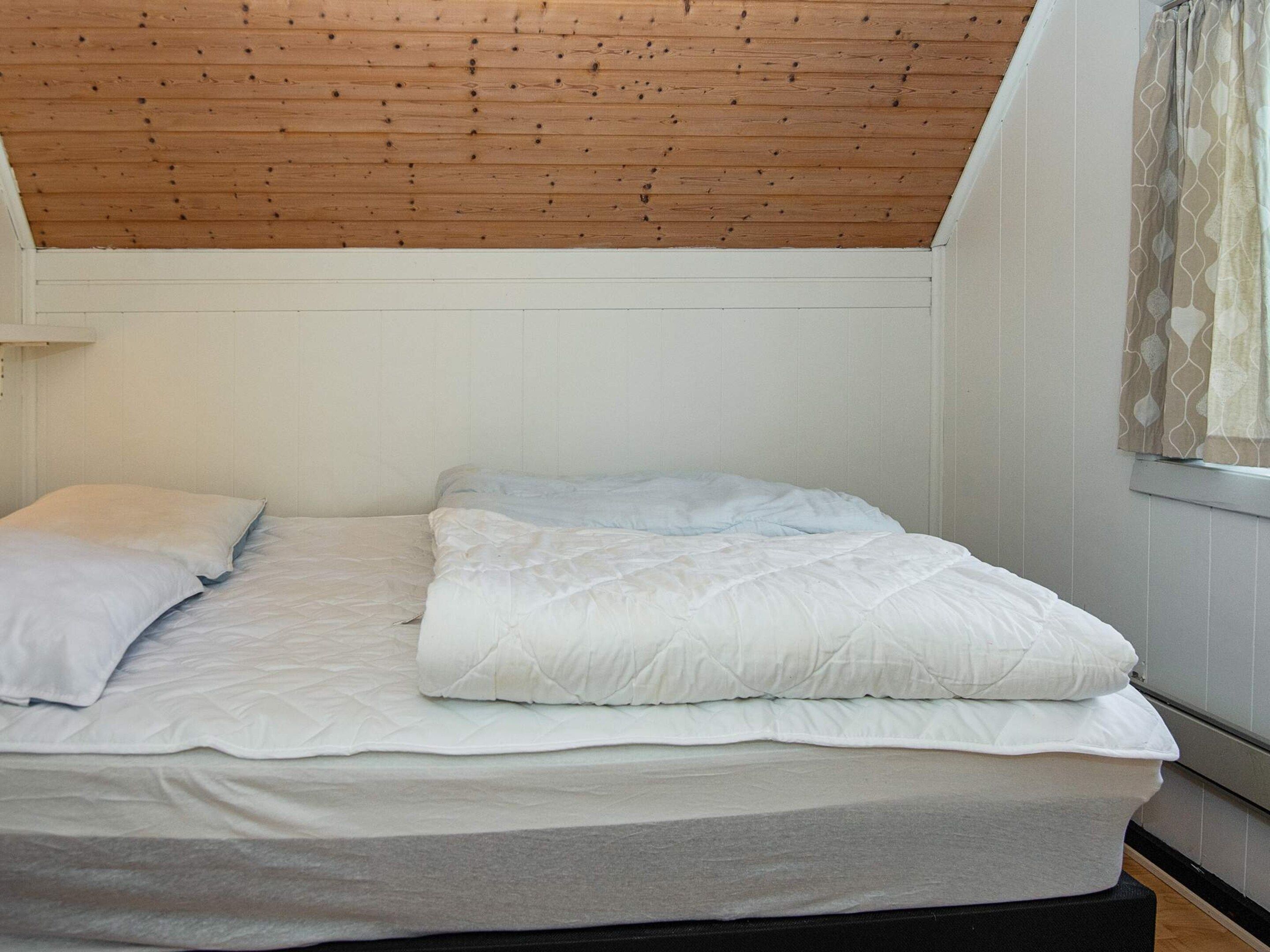 Una cama individual con ropa de cama blanca en una habitación con vigas de madera en el techo y paredes blancas.