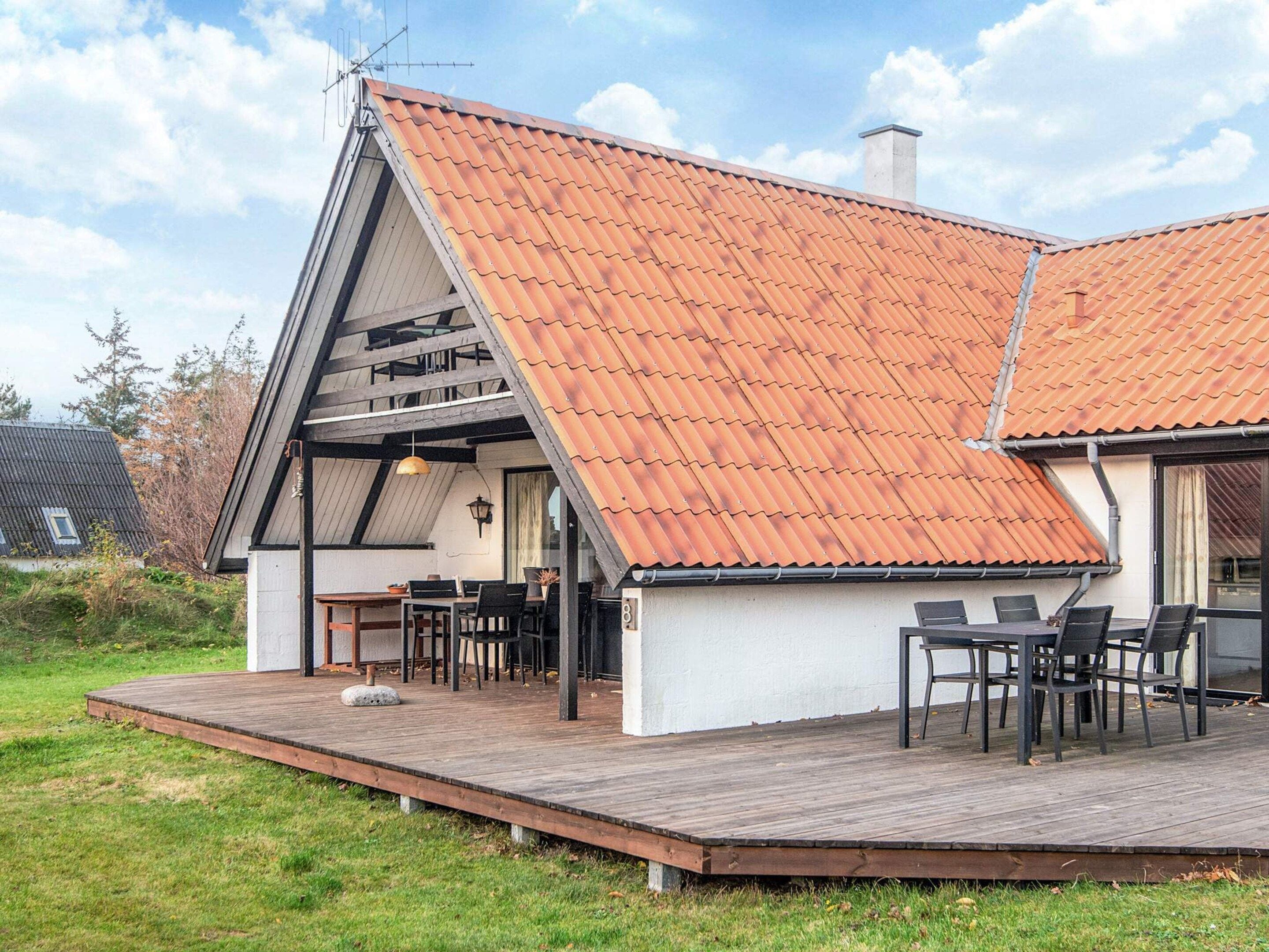 Una casa moderna con techo inclinado, una terraza de madera y un comedor con mobiliario exterior.