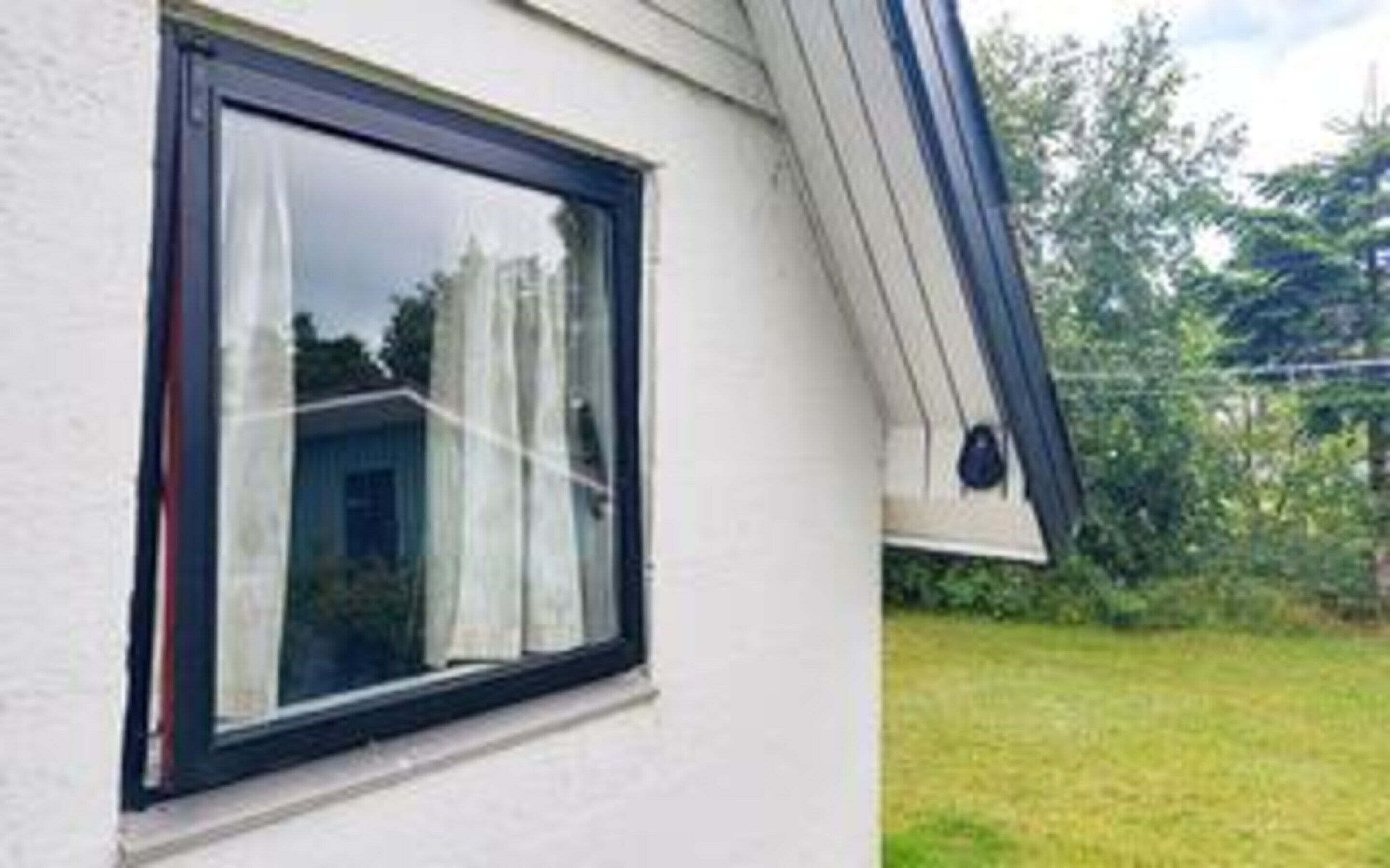 A window with a black frame and white curtains, a white wall, and a grassy yard with trees.