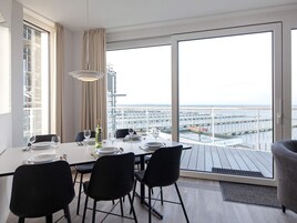 A dining area with a table set for four, large windows with a view of the sea and a pier, and a balcony with a view.