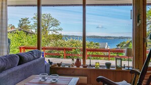 Quarto com sofá, mesa de madeira com copos, uma cadeira e vista para o mar através de amplas janelas.