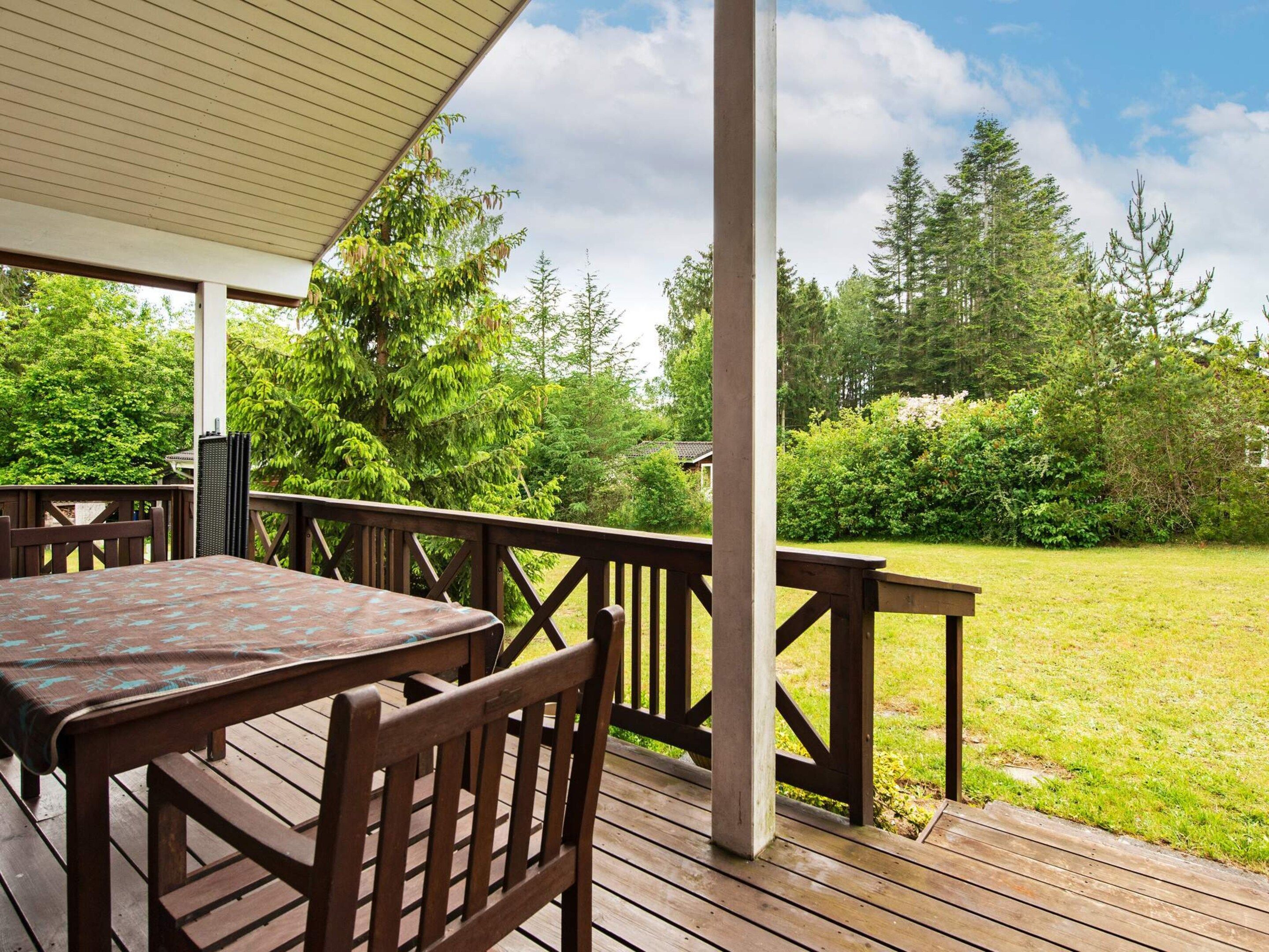 A covered outdoor deck with a table and chairs, surrounded by a grassy area and trees.