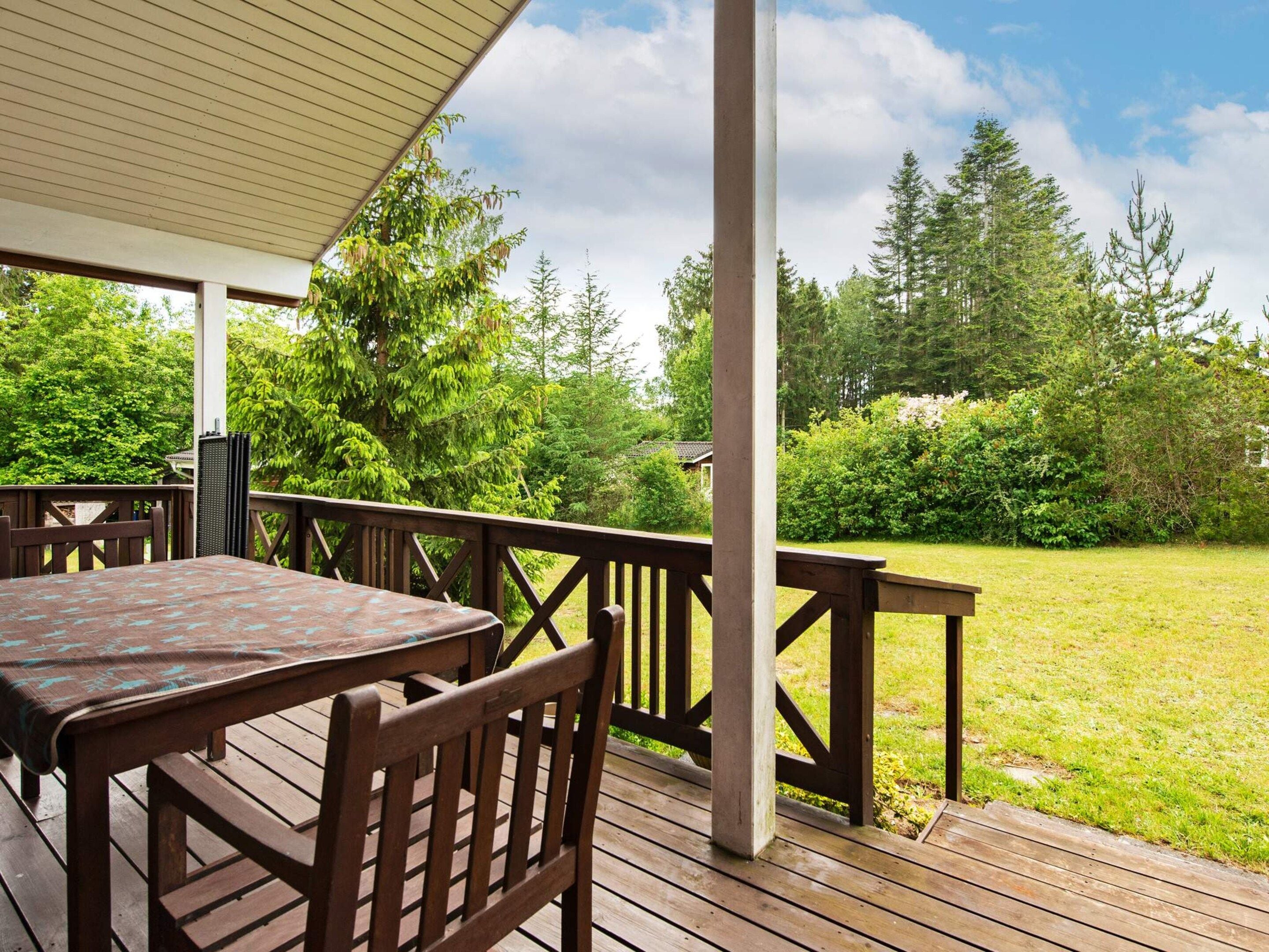 Een overdekt terras buiten met een tafel en stoelen, omringd door een gazon en bomen.
