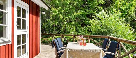 Una terraza de madera con mesa y sillas, un edificio rojo con detalles blancos y un paisaje boscoso al fondo.