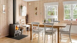 A room with a wood-burning stove, a dining table with chairs, and a window with a view of trees. - 8 Person Holiday Home in Skanes Fagerhult (Skånes Fagerhult)
