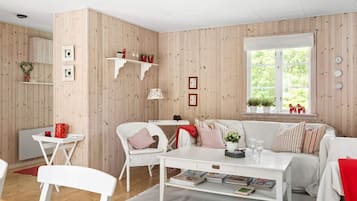 A cozy living room with a white sofa, a dining table, and a coffee table. There are shelves with decorative items and a window with a view of greenery.
