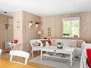 A cozy living room with a white sofa, a dining table, and a coffee table. There are shelves with decorative items and a window with a view of greenery. - 8 Person Holiday Home in Skanes Fagerhult (Skånes Fagerhult)