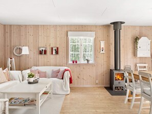 A cozy living room with a sofa, a fireplace, a dining table with chairs, and a window with a view of trees. - 8 Person Holiday Home in Skanes Fagerhult (Skånes Fagerhult)