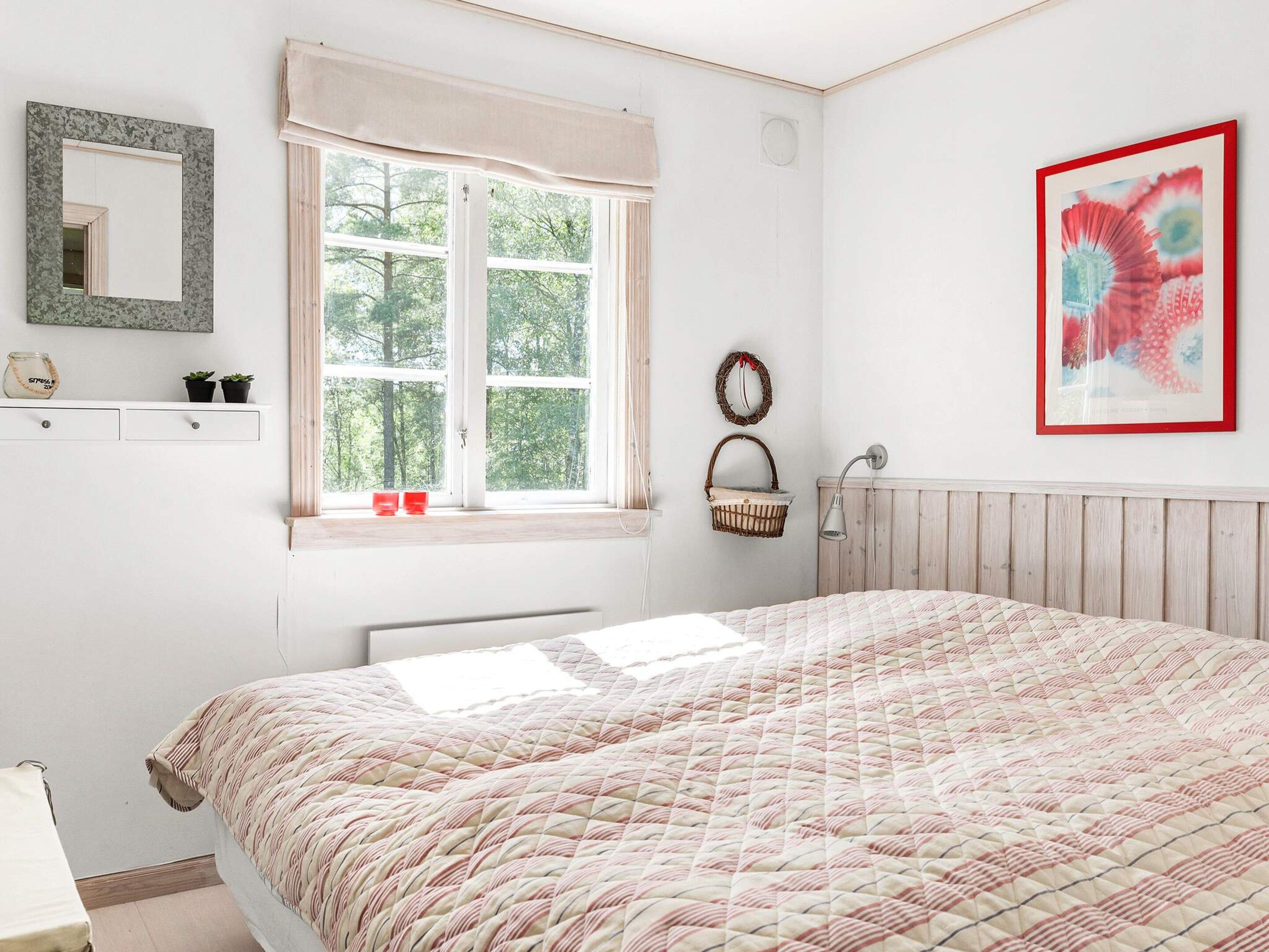 Un dormitorio con una cama grande, una ventana con cortinas, una lámpara de noche, un espejo montado en la pared y un cuadro enmarcado.
