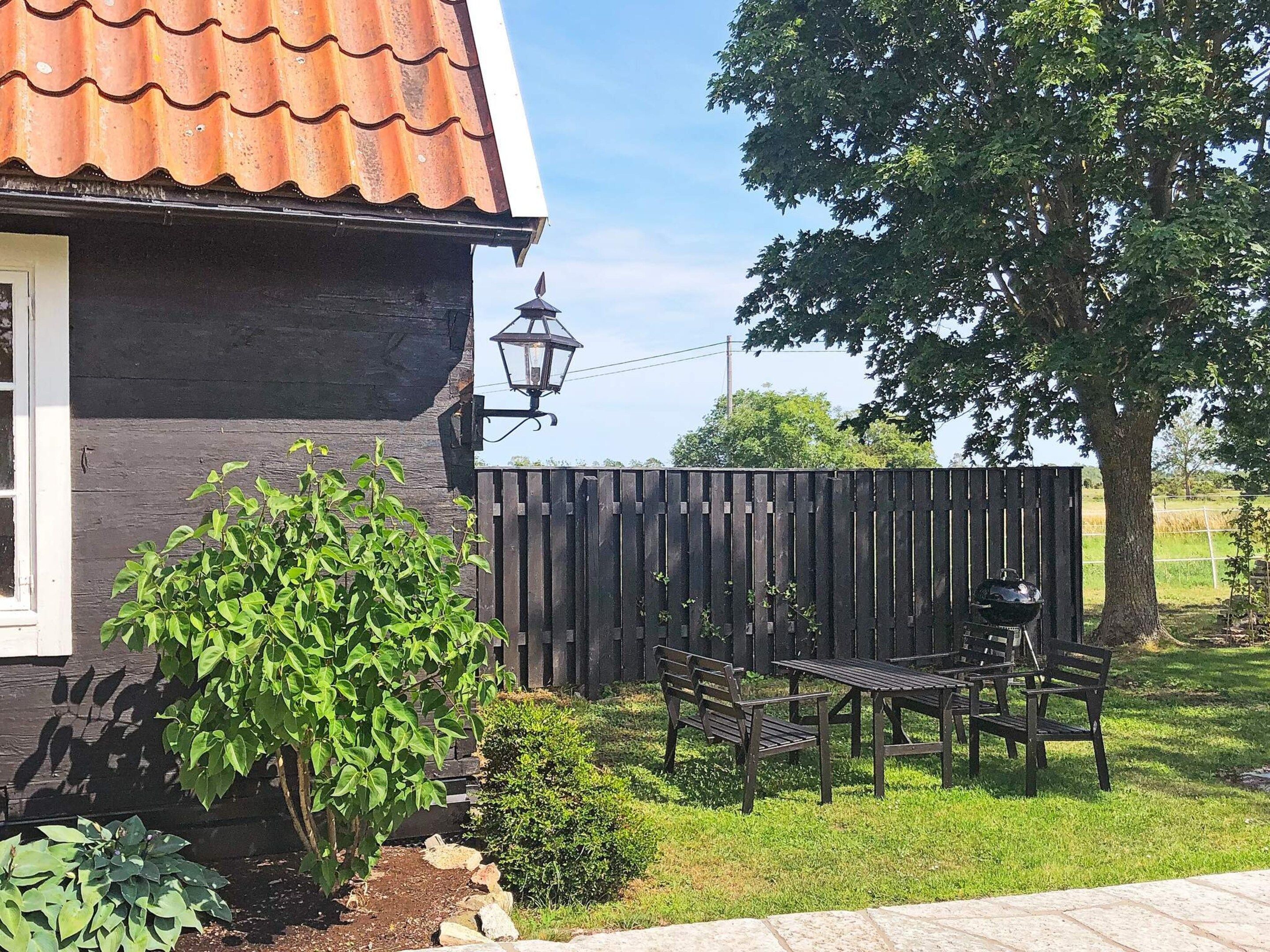 A house with a black exterior and a red-tiled roof, a wooden fence, a bench, a table, and a grill.
