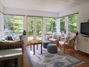 A sunlit room with a sofa, ottomans, and wicker chairs, a television, and large windows with a view of greenery. - 4 Person Holiday Home in Frederiksvaerk (Frederiksvaerk)