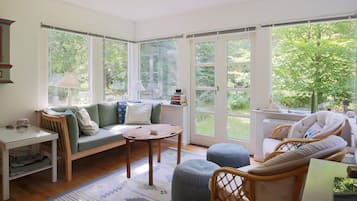 A bright room with a sofa, ottomans, and a table. There are large windows and a view of greenery outside.