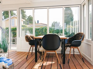 A sunlit room with a wooden table and chairs, large windows, and a view of a garden and buildings outside.