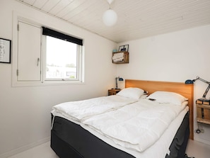 En dobbeltseng med trægavl, to sengeborde med lamper, et vindue med mørklægningsgardin og en vægmonteret hylde med håndklædestang.