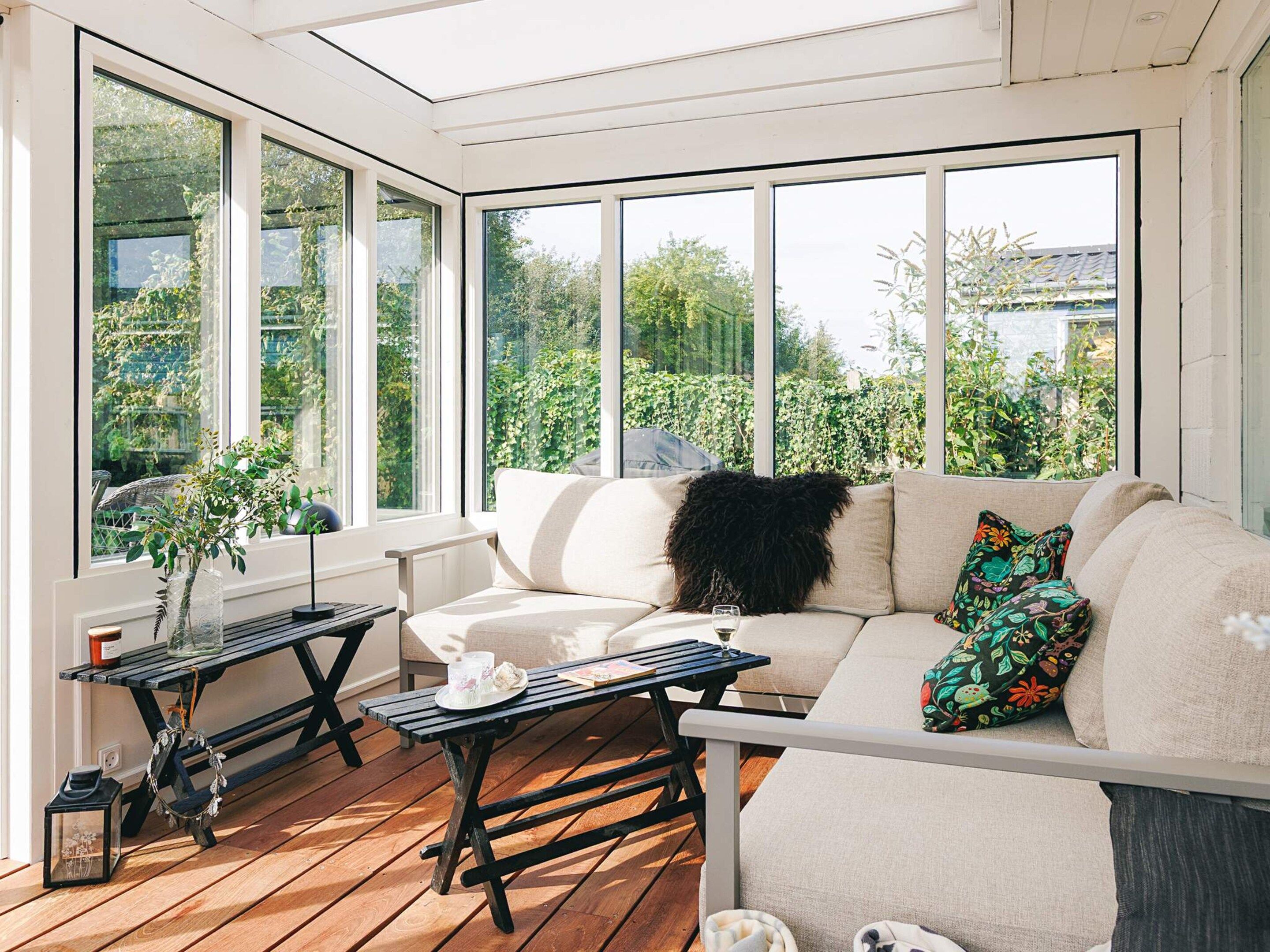 A sunroom with a wooden deck, a sofa with cushions, a table with a glass top, and a vase with greenery.