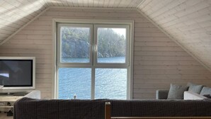 A room with a sloped ceiling, a large window overlooking a body of water and greenery, a television, and a checkered sofa. - 6 Person Holiday Home in Rubbestadneset (Bomlo)