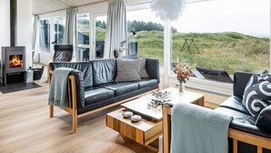 Een moderne woonkamer met een open haard, leren bank, houten salontafel en grote ramen met uitzicht op de natuur.