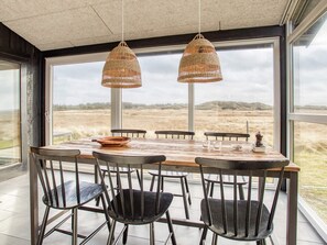 An indoor dining area with a large table and chairs, two hanging wicker lamps, and a view of a natural landscape through large windows.