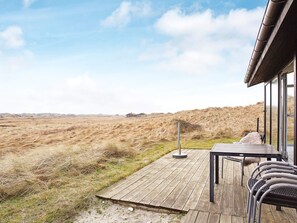 Une terrasse en bois avec une table et des chaises, offrant une vue sur un paysage verdoyant, avec des dunes et un ciel dégagé.
