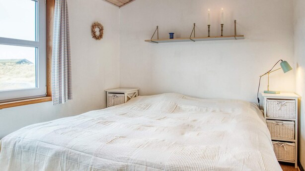 A bedroom with a large bed, a window with curtains, a wall-mounted shelf with candles, and a bedside table with a lamp.