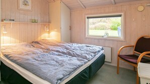 A bedroom with a bed, a chair, a window, and a shelf with books and bottles. - Tranquil Oasis in Blavand-by Traum (Rudkobing)